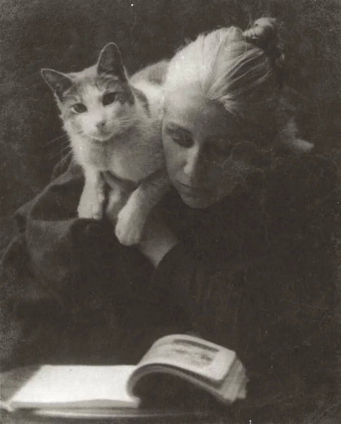 A black and white vintage photo of a woman holding a cat while reading a book