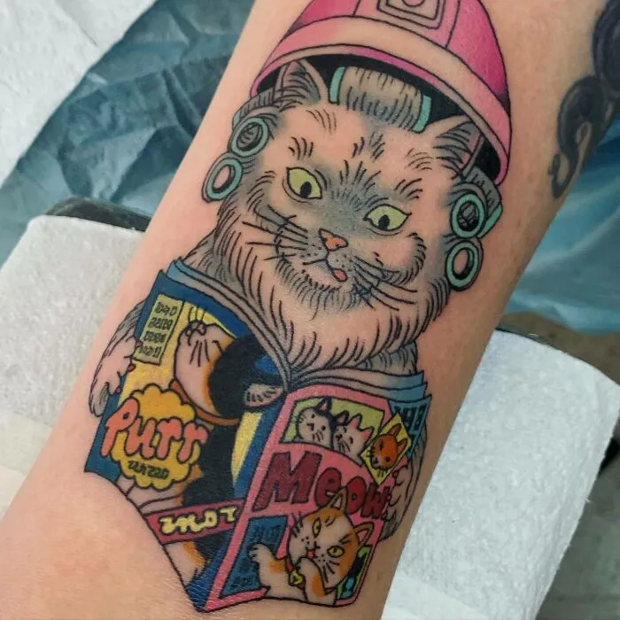 Creative cat tattoo on an arm, showing a cat with a pink hair dryer reading a magazine about cats.
