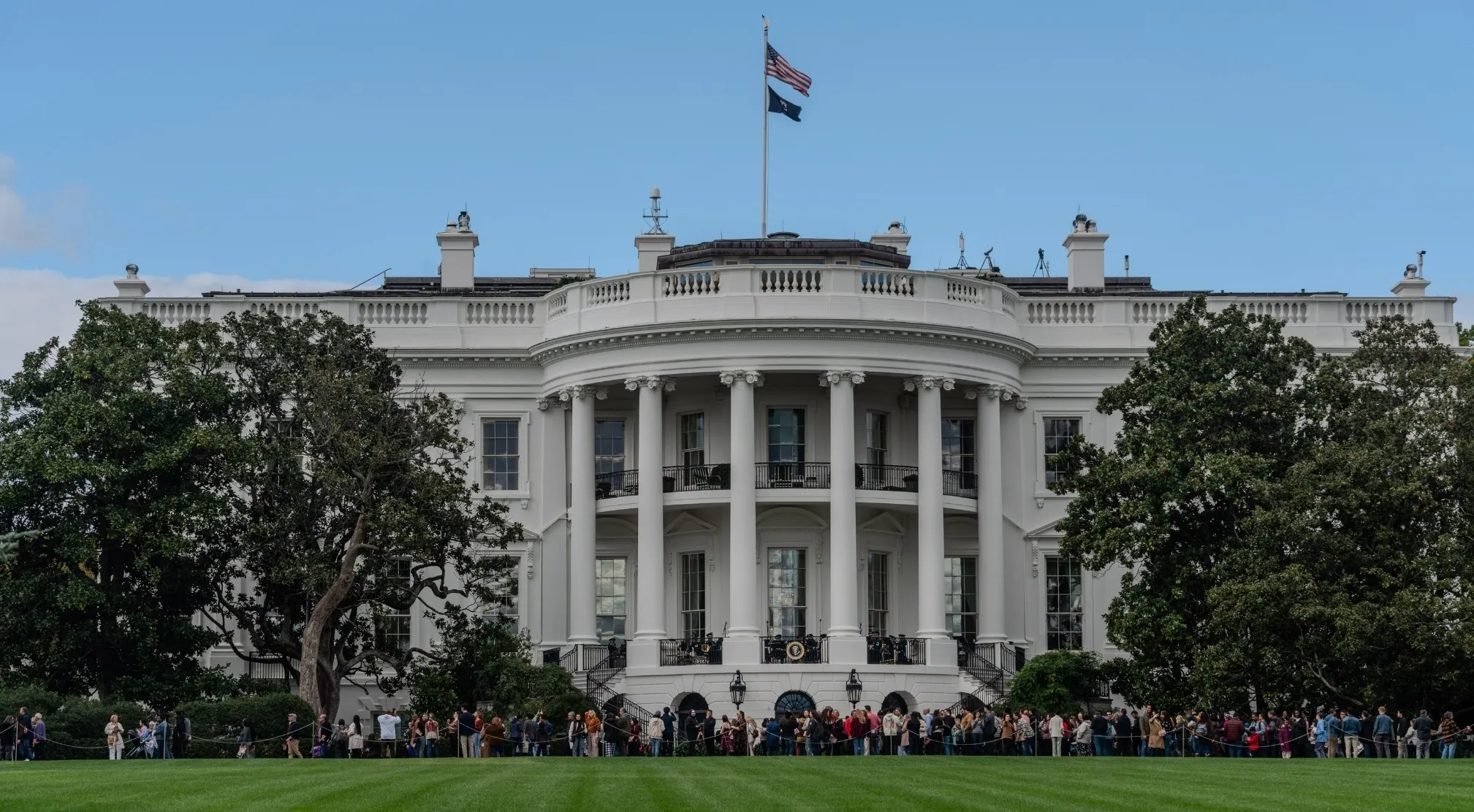 The White House, a US President residence, with a crowd of people and trees in front, prompts UFO and Conspiracy Theories Trivia.