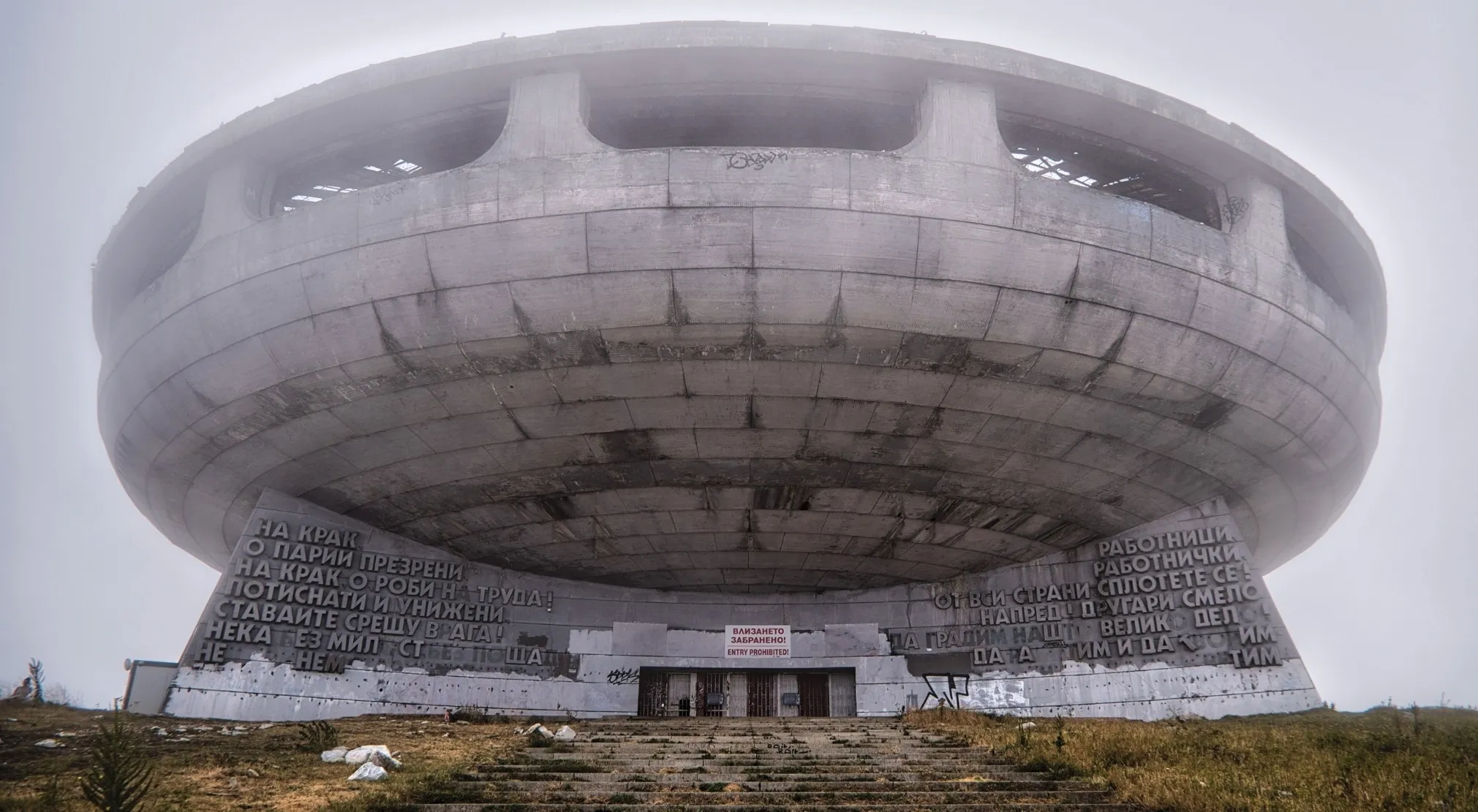 Mist shrouds the UFO-like Buzludzha Monument in Bulgaria, a concrete conspiracy theories structure with Cyrillic text and an entry prohibited sign.