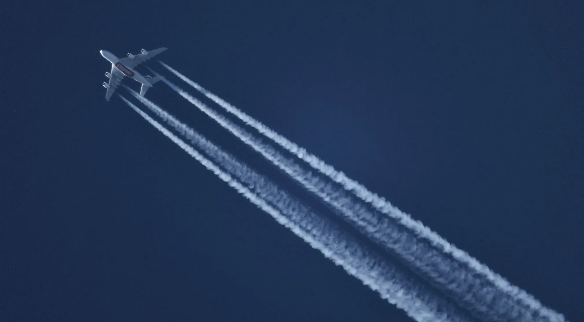 A jet streaks across a dark blue sky, leaving four white contrails, often mistaken for chemtrails. UFO and conspiracy theories.