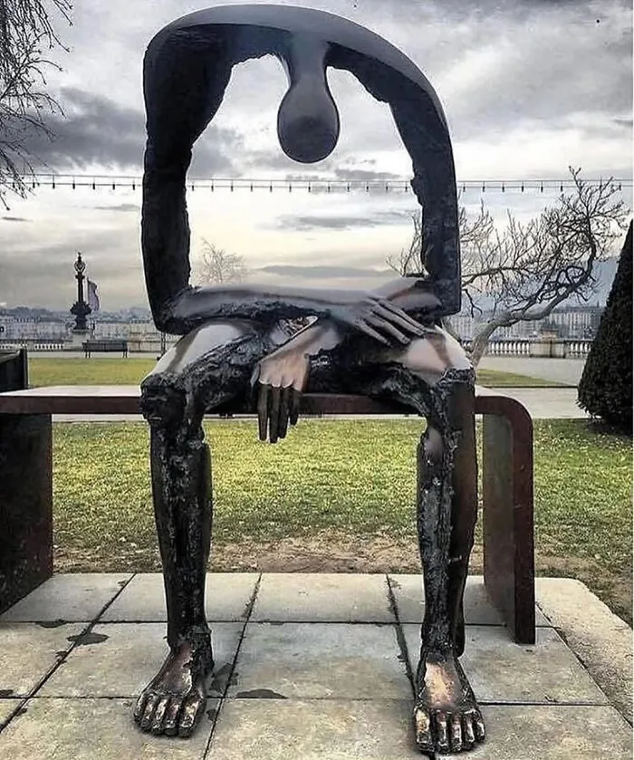 A unique bronze street art sculpture of a figure sitting on a bench, head bowed. A stunning example of street art.