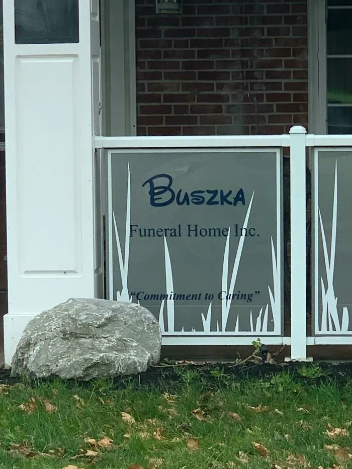 A sign for Buszka Funeral Home Inc. with the commitment to caring motto, a humorous ad placement next to a large grey rock.