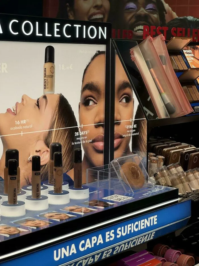 A makeup display with various foundation bottles and a large ad with hilarious ad placements, showing a reflection of a woman.