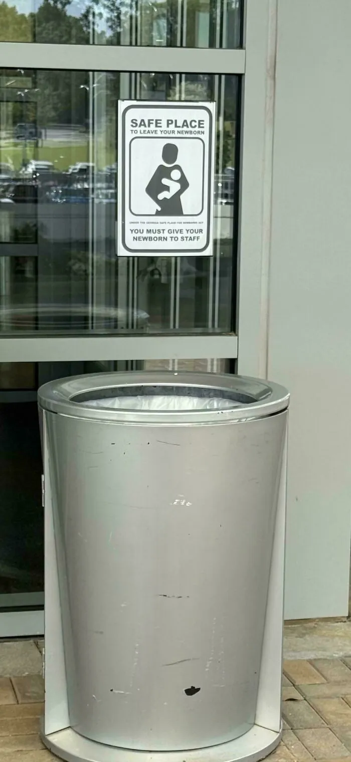 A sign on a glass door that reads SAFE PLACE TO LEAVE YOUR NEWBORN placed above a metal trash can, showcasing hilarious ad placements.