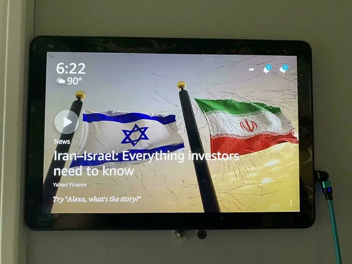 A cracked tablet screen displaying a news headline about Iran-Israel relations, symbolizing a dystopian nightmare amidst the American Dream.