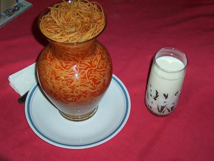 Unhinged images: A glass vase filled with spaghetti and sauce, served on a plate next to a glass of milk.