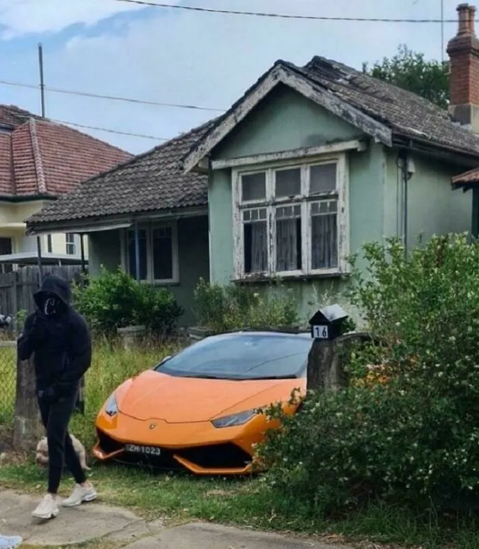 An orange Lamborghini parked by a run-down house, creating an unhinged image.