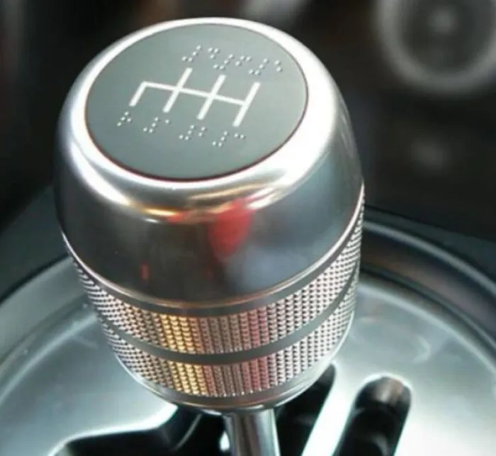 A close-up of a silver car gear shift knob with braille markings, one of many unhinged images.