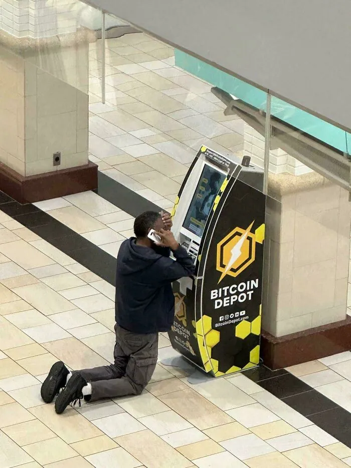 A man kneeling and talking on his phone in front of a Bitcoin Depot ATM, an unhinged image.