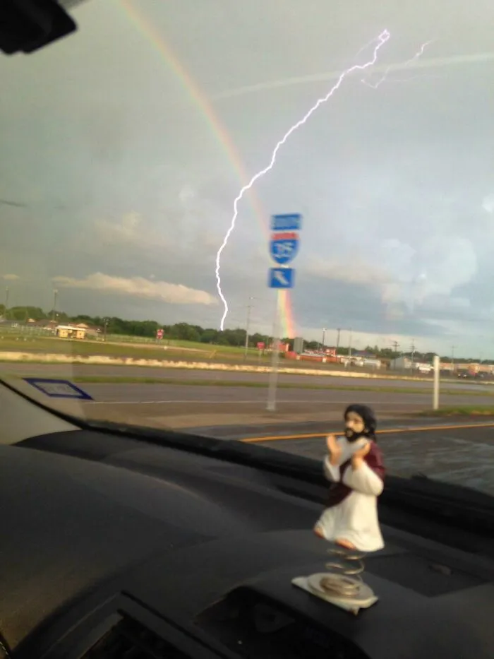 A bobblehead Jesus on a car dashboard, with a rainbow and lightning strike outside, a moment synced just right.