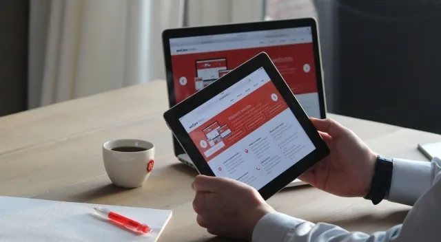 A person viewing a red and white website on a tablet, with a laptop, coffee, and pen nearby. General Knowledge Quiz.
