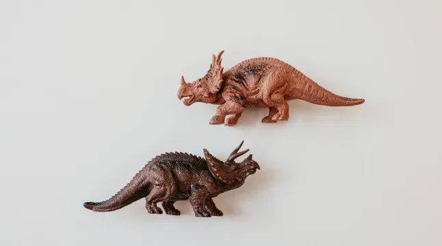 Two toy dinosaur figures, one brown and one darker brown, posed on a white background for a general knowledge quiz.