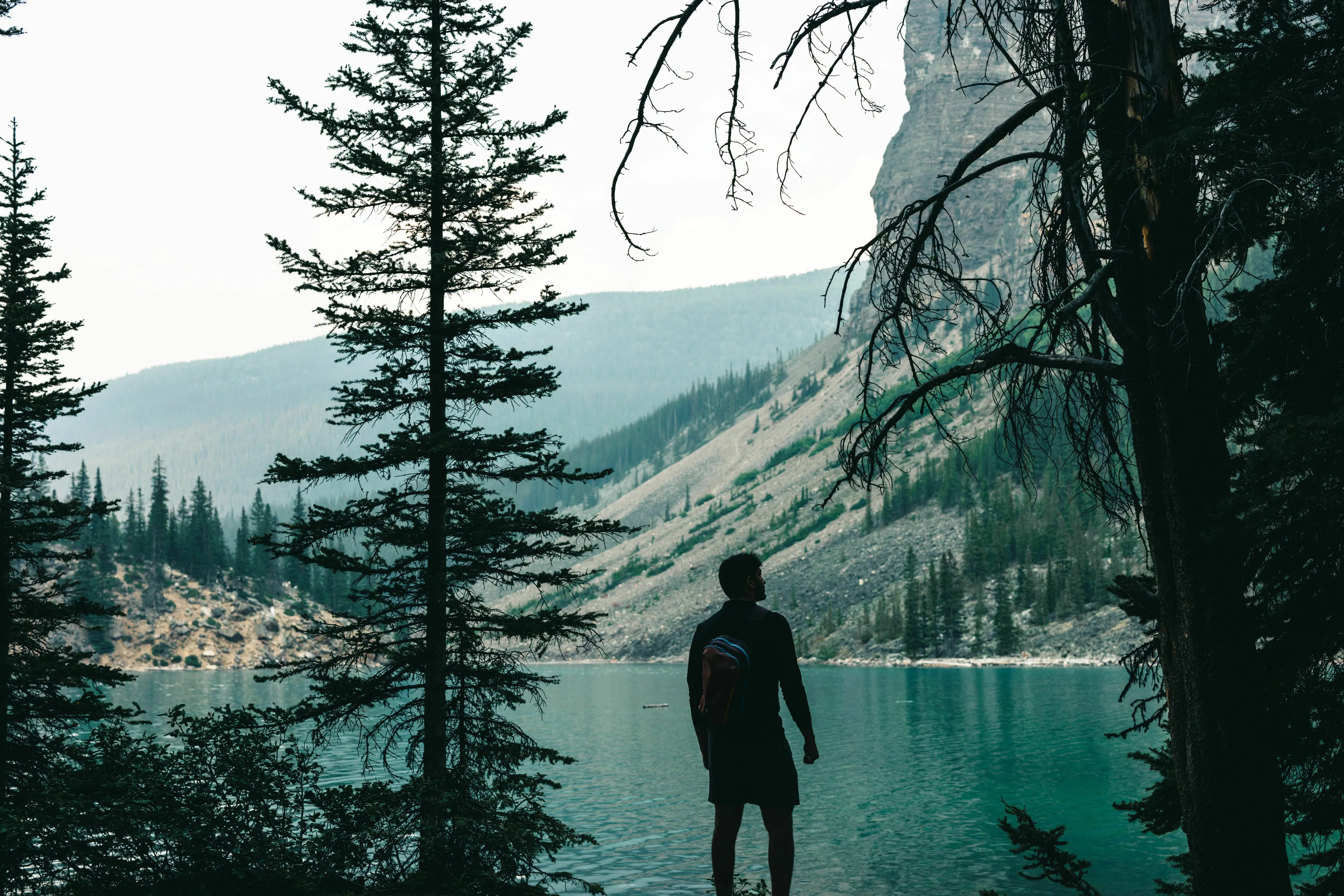 A person standing by a natural lake surrounded by pine trees and mountains, evoking a sense of adventure and nature.