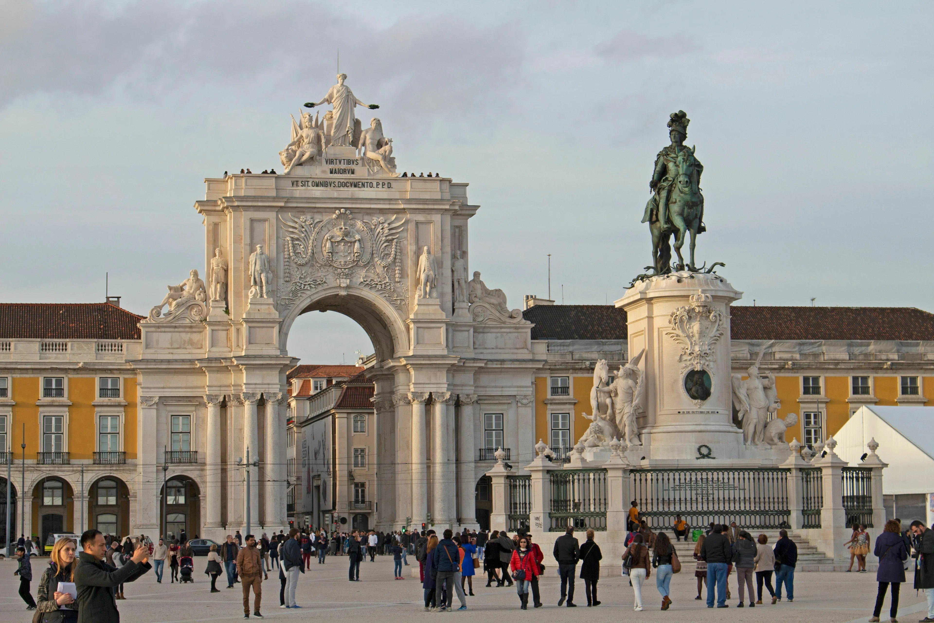 People gather in Lisbon, Portugal, in a grand square with a large triumphal arch and a statue.