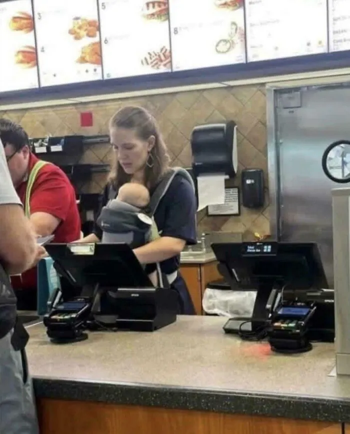 A woman working at a fast-food counter with a baby in a carrier, illustrating USA dystopia.