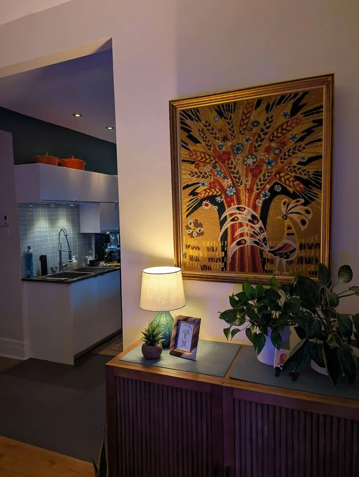 An embroidered framed picture of a bird next to a table lamp, great thrift store finds.