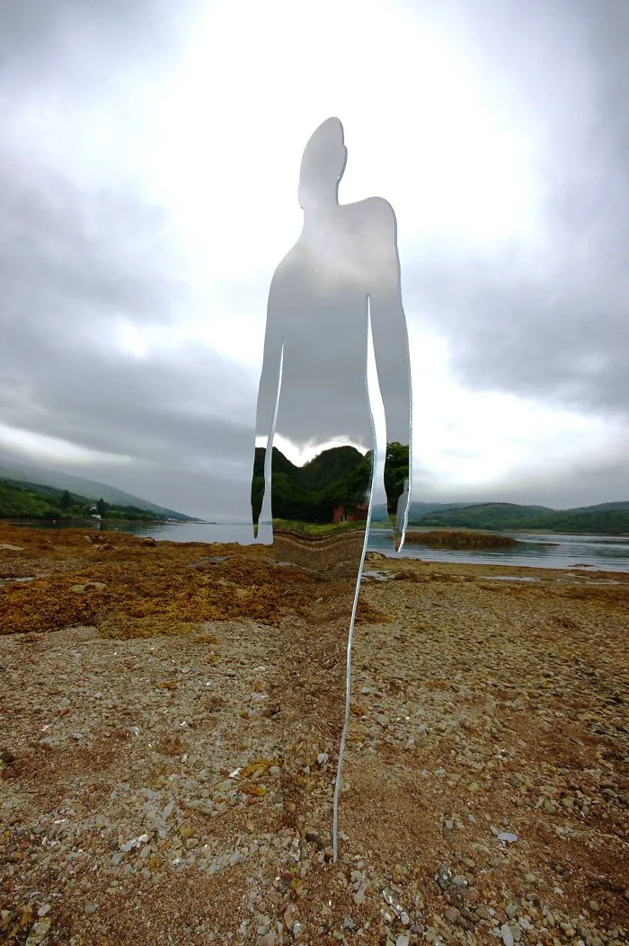 Reflective standing figure sculpture blending into rocky shoreline scenery