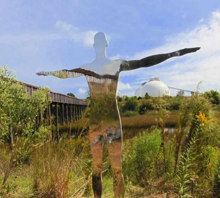 Reflective sculpture with outstretched arms in a grassy area with a bridge and dome in background
