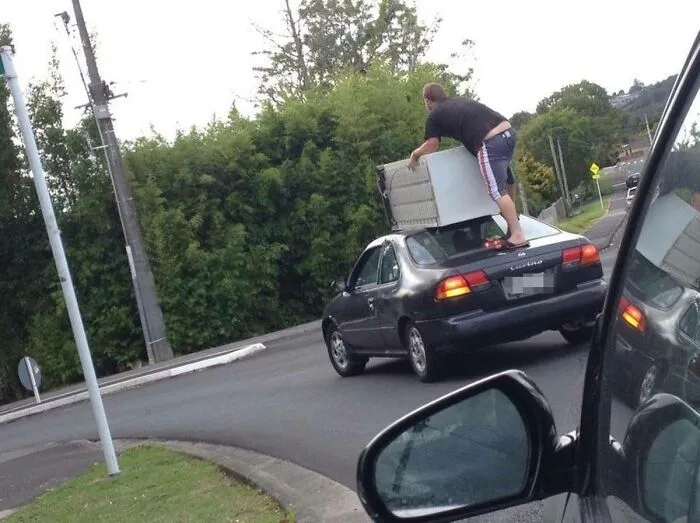 Man precariously balancing large appliance on car roof in moving fails