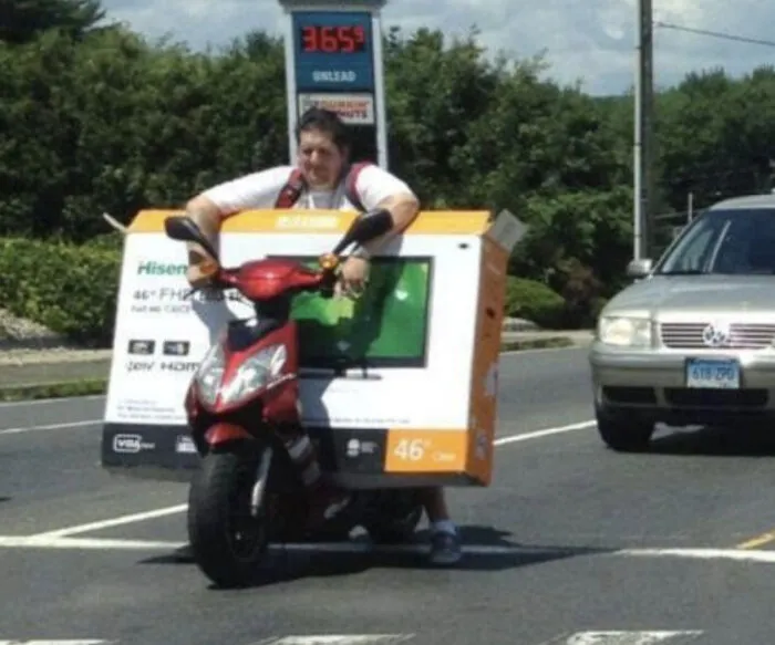 Moving fails photo of person awkwardly carrying large TV box while riding scooter