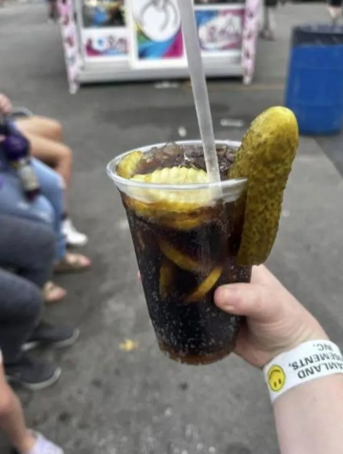 A hand holding a cup of dark cola with pickles inside, representing weird food combinations