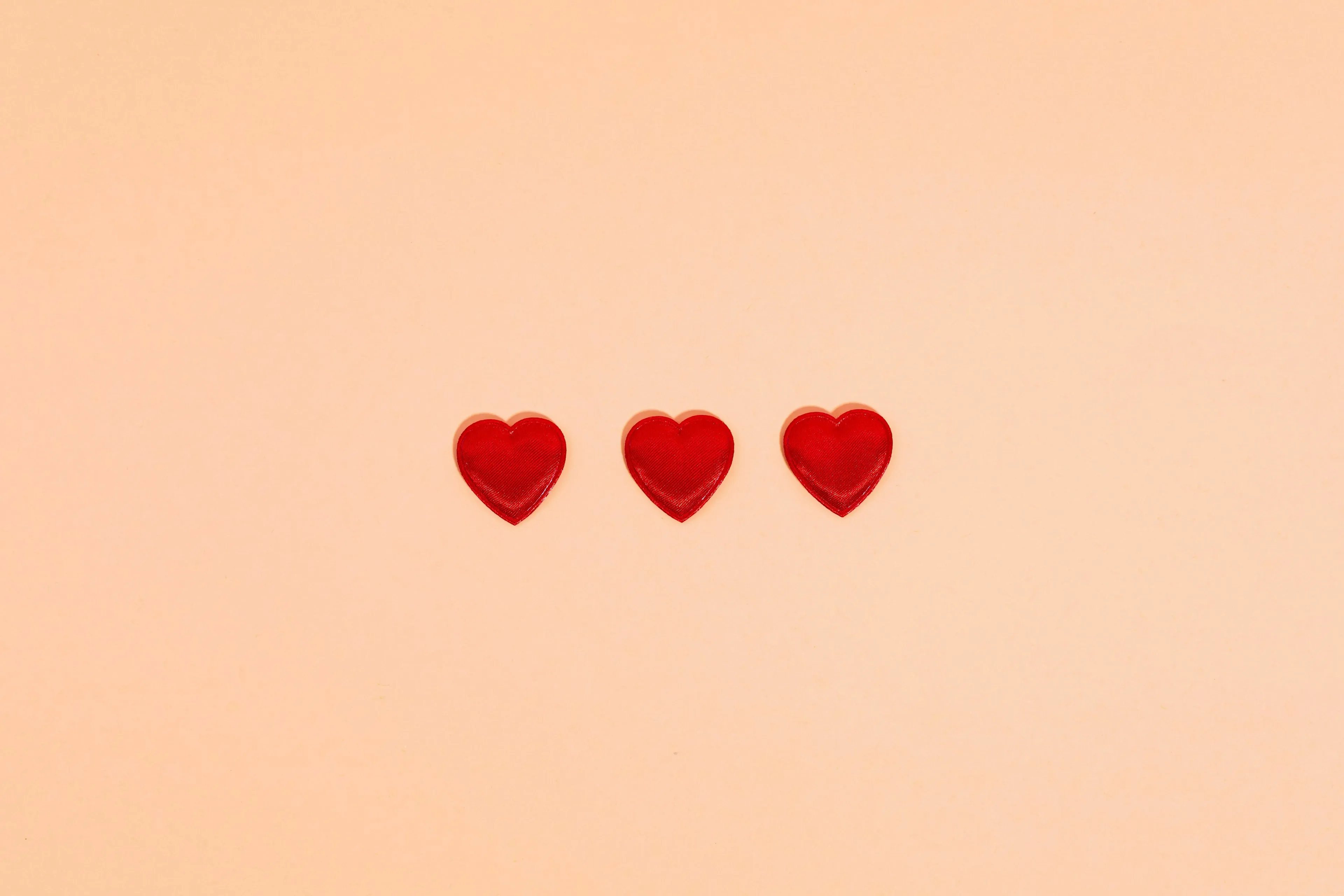 Three small, identical red heart shapes aligned horizontally on a soft peach background.