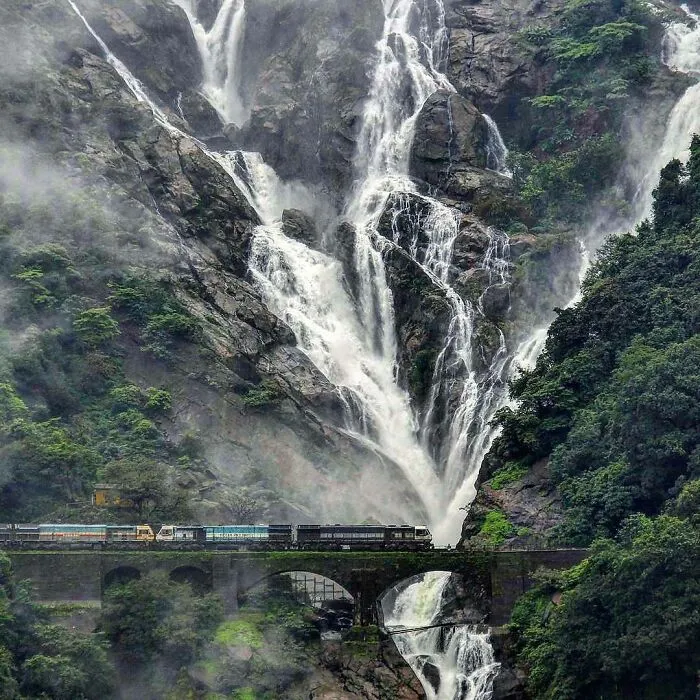 A train passing over a stone arch bridge with a massive waterfall in the background, a beautiful example of infrastructure