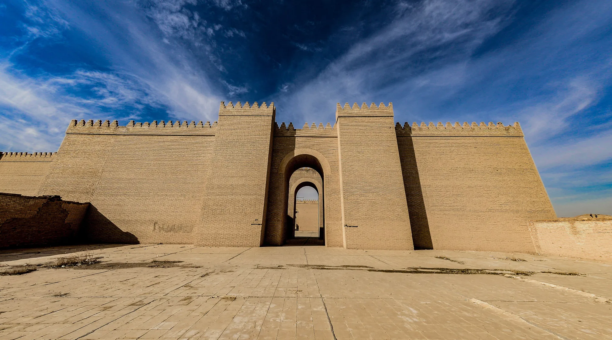 The grand ancient stone walls and entrance gate of the historic city of Babylon under a bright blue sky.