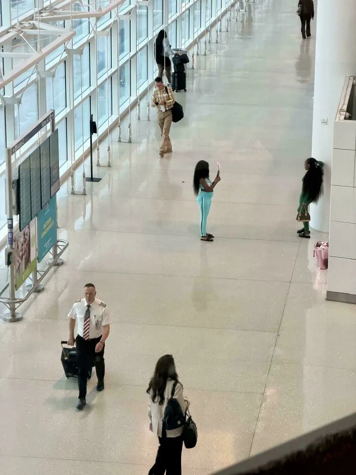 Two women blocking a wide airport hallway to take photos, showcasing main character vibes.