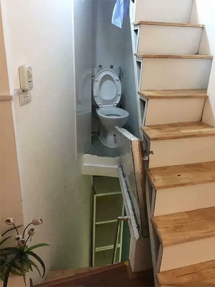 A cramped toilet situated awkwardly under a wooden staircase in a poorly designed home