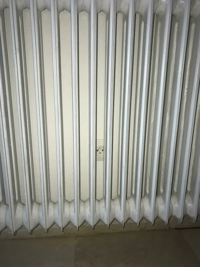 An electrical outlet hidden behind a white wall radiator in a poorly designed house