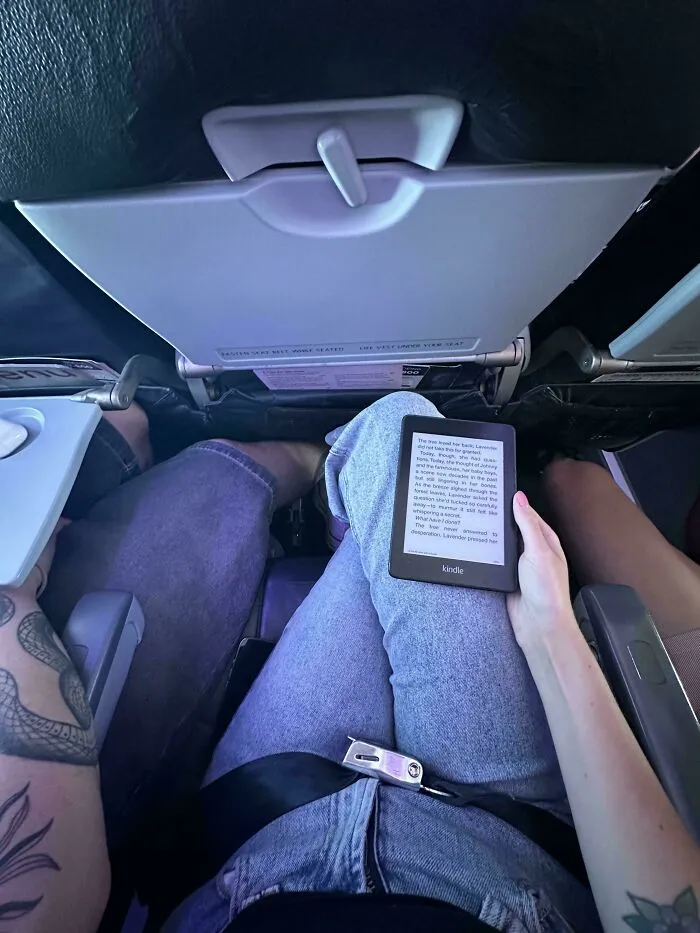 Person holding a Kindle over their seatmate in a cramped airplane, demonstrating main character lives behavior.