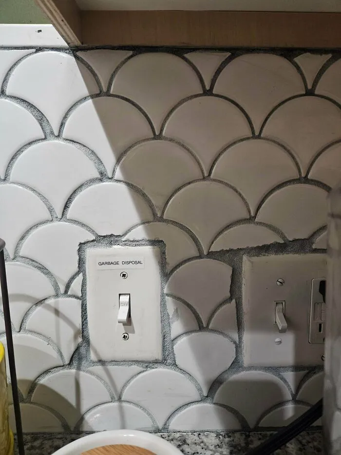 A poorly installed fish scale tile backsplash fitting awkwardly around light switches in houses with poor planning.