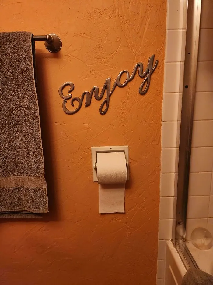 An orange bathroom wall featuring an Enjoy sign above a toilet paper roll, showing houses with a big vision.