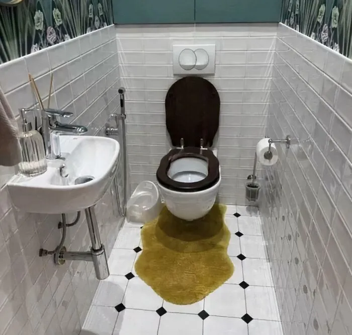 A narrow bathroom with white tiled walls and a yellow bath mat shaped like a spilled liquid under the toilet.