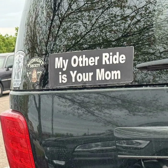 A black car rear window featuring a wild bumper sticker that says, My Other Ride is Your Mom.