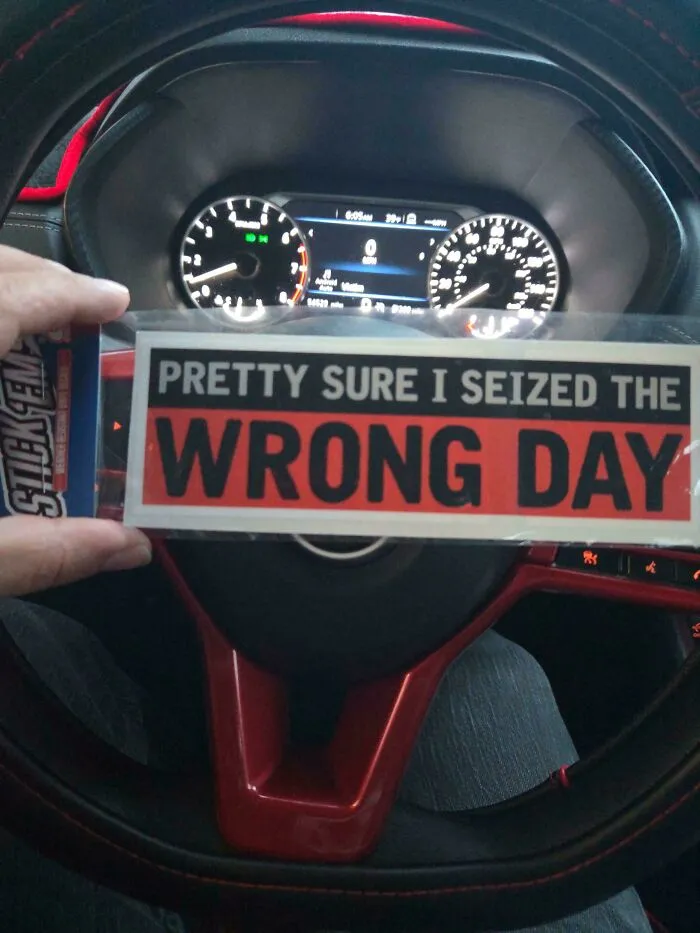 A hand holding a wild bumper sticker reading, Pretty Sure I Seized The Wrong Day, inside a car.