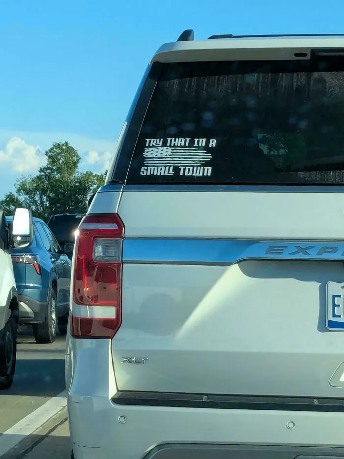 A white SUV displaying wild bumper stickers on the back glass reading Try That In A Small Town with a flag graphic.