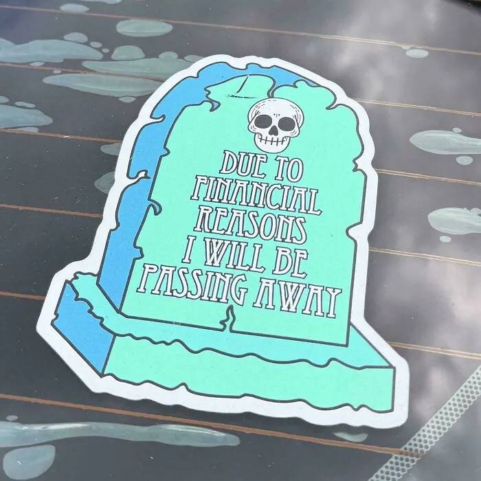 Funny tombstone bumper stickers on a car window with humorous text about finances.