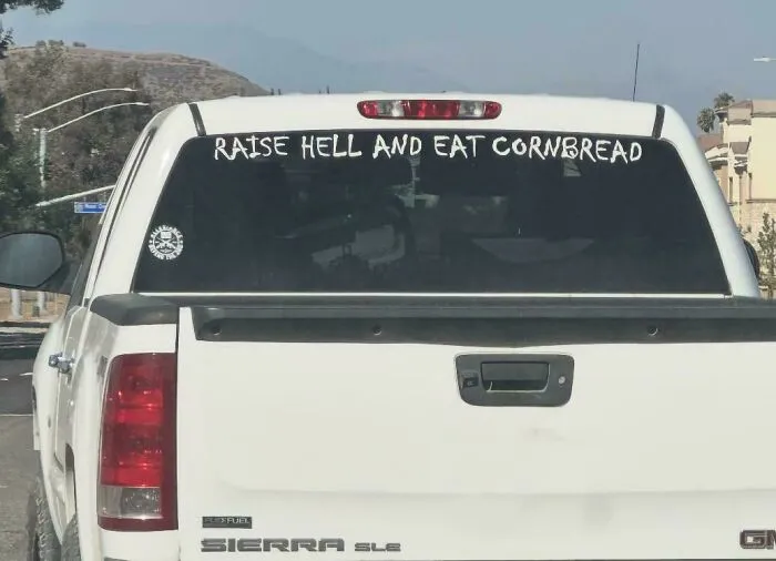 White truck with bumper stickers decal saying Raise Hell And Eat Cornbread on the rear window.