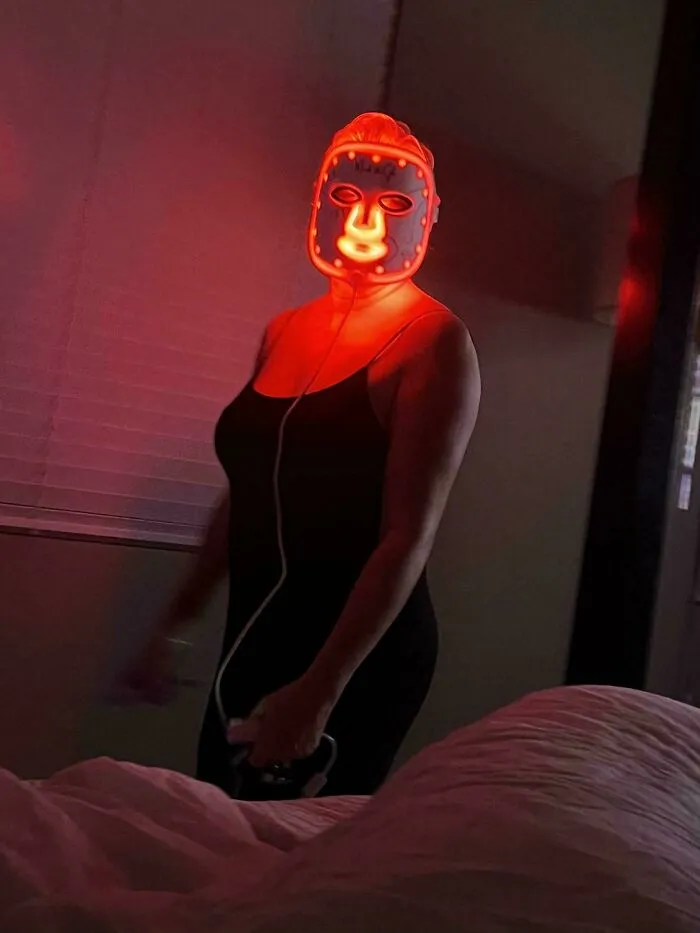 A woman standing in a dark room wearing a glowing red LED skincare mask, illustrating unhinged things wives do.