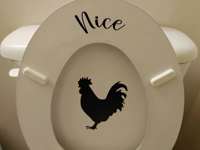 A white toilet seat with a black rooster decal underneath a sticker that reads Nice