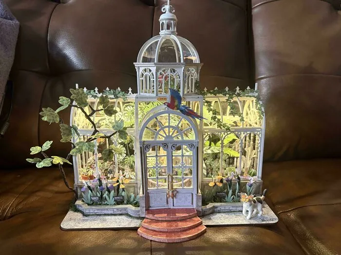 A glass greenhouse book nook miniature featuring a parrot and green plants inside