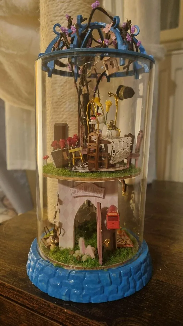 A whimsical book nook enclosed in a glass dome featuring a miniature tea party setup with tiny chairs