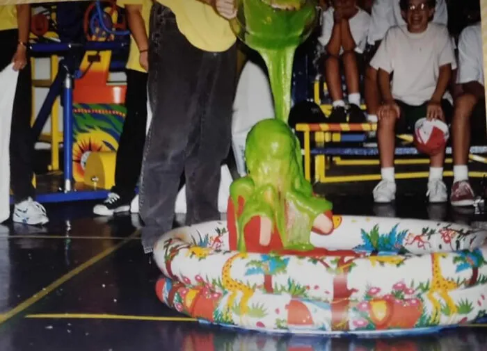 A 1990s television studio scene showing green slime being poured over someone in an inflatable pool