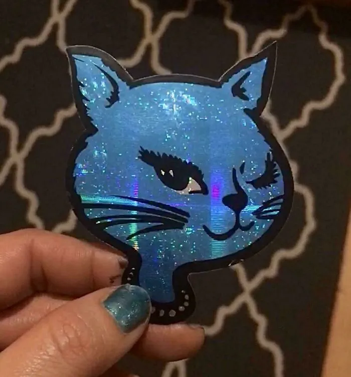 A hand holding a sparkling, holographic blue cat sticker from the 90s with a winking face
