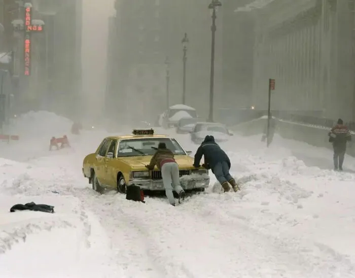 A yellow cab being pushed through heavy snow in 90s New York City