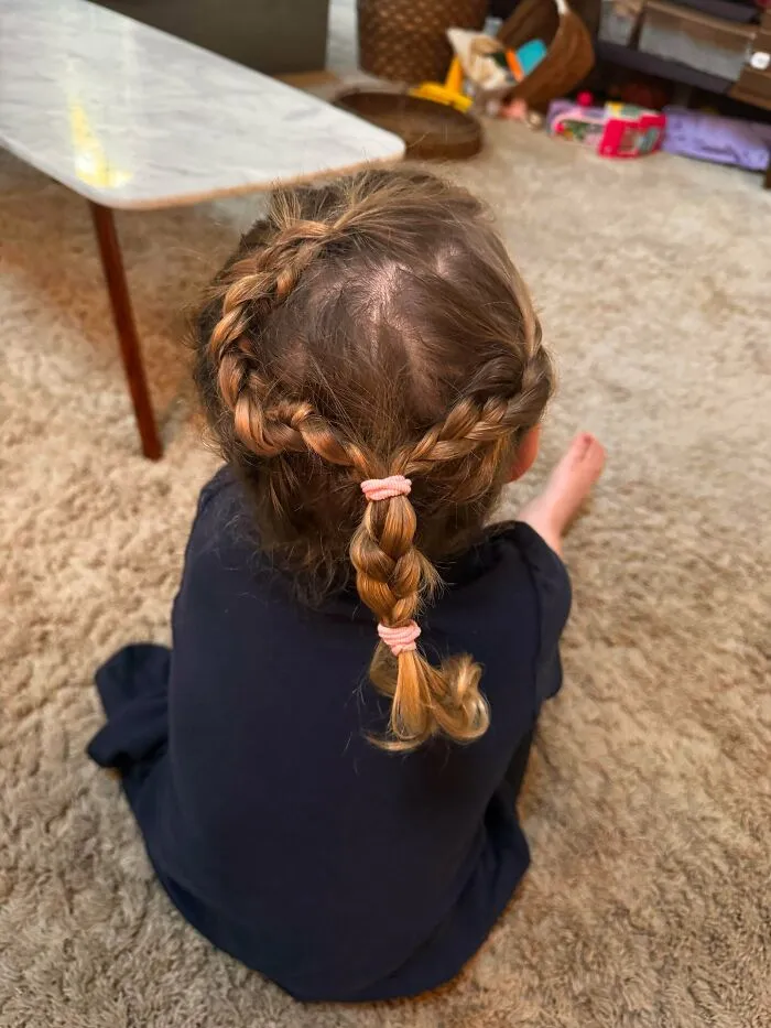 A young child sitting on the floor displaying a neatly styled braided ponytail created by their parent.