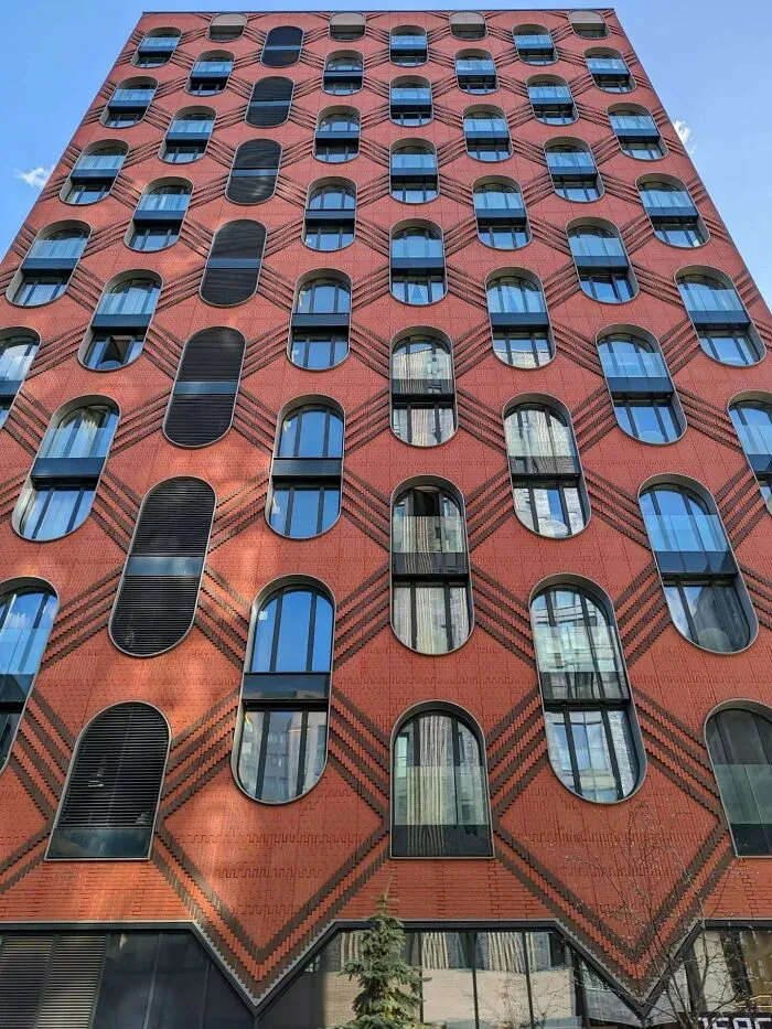 Low angle view of a tall red brick facade with rounded windows, featuring modern architectural masterpieces.